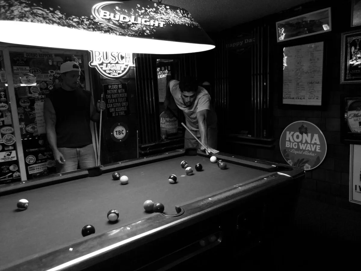 Eddy lining up a shot on a pool table in a bar, in black and white, a friend waiting with a cue.